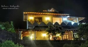 A two-story stone house illuminated at night, with a wooden roof, balcony railing, and surrounding trees