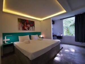 A modern bedroom with a bed, teal headboard, nightstand, lamp, abstract painting, ceiling lights, concrete floor, large window with palm trees, and a small sink area.