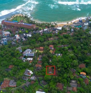 Aerial view of Hikkaduwa town with houses and trees, near the beach with waves, and a red square marking a specific property.