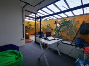 A cozy workspace with a white desk, laptop, and desk lamp, surrounded by green plants and yellow walls. The room features a glass ceiling, bean bags in green and blue, and an open door leading to another area.