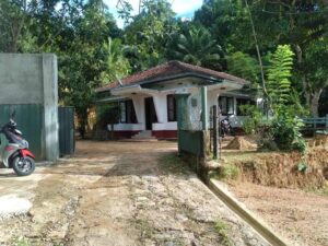 Front view of a red-tiled house with open gate, scooter, tropical greenery, and unpaved yard.