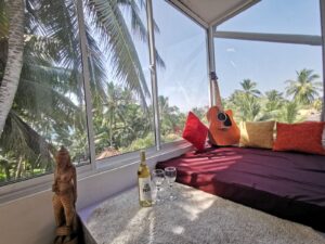 A cozy lounge with a bed, red and yellow pillows, a guitar, wine bottle, and glasses, next to a large window with a tropical palm tree view. A wooden statue stands nearby.