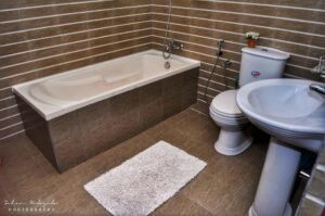 A bathroom with a white bathtub, toilet, sink, brown tiled walls, and a fluffy white mat on the floor.