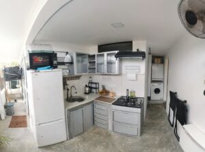 A small kitchen with a refrigerator, microwave, sink, stove, cabinets, and a washing machine, featuring a ceiling fan and some fruit on the counter.