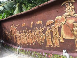 Wall featuring a detailed relief sculpture of people, an elephant, and umbrellas, with a tiled roof and colorful floral base, surrounded by trees.