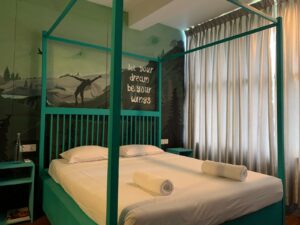 A bedroom with a teal canopy bed, white linens, rolled towels, and a nightstand. The wall features a mural of a landscape with the text "let your dream be your wings."