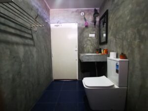A small bathroom with gray concrete walls, a white toilet, concrete sink, black-framed mirror, towel rack, and blue tile floor.