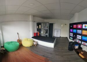 A room with a bed, green and yellow bean bags, a guitar, a bookshelf, and a large TV displaying streaming apps, on a wooden floor.