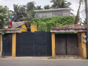 A house with a tiled roof, yellow and black gate, surrounded by palm trees and green vines, featuring a sign and decorative mailboxes.