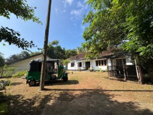 White house with a tiled roof, surrounded by trees, and a green auto rickshaw parked in the yard under a clear blue sky.